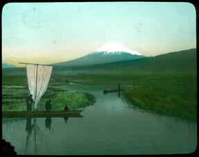 People in boats on waterways through rice fields; snow-covered mountain in background.