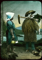 Two people in costume posing in front of painted backdrop - one in grass coat and hat, carrying farm implement over shoulder and holding vegetables; the other, a woman in a blue dress with a red (over mantel), legs bare to the knees, wearing a white kerch