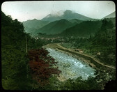 River with manmade embankment and road on one side; line of buildings along embankment; forest on other side of river, town and mountains in background.