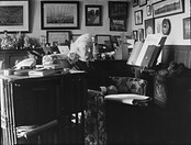 Interior of cluttered room, Augusta Crofton Dillon seated on armchair, surrounded by ornaments. Many paintings and photographs on wall, also shows reading stand with garden magazine and two books.