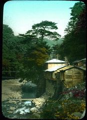 Buildings built up on stone walled riverbank with trees in background.