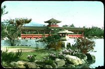 Long red wooden covered bridge with ornamental roof; woods on either side of bridge, woods and mountain behind; water and landscaped point of land with flowers in foreground.