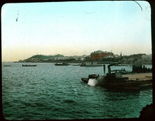 Harbour of city; jetty with steamboats in foreground; other boats and buildings in background.