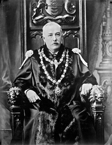 A distinguished gentleman in robes and chain of office sitting on a throne (of sorts)