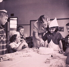 Woman helping young girl in a paper hat slice a birthday cake at a party while other children look on