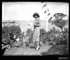 Woman with clutch bag and hat posing for the camera with a regatta in the background, 1920-1935