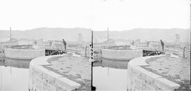 Navigation bridge (iron) on curved lock, dock on canal entrance, another bridge, towns old mill, hills, in background