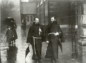 "Two unidentified Franciscan friars walking alongside a church carrying an umbrella" is Fathers Bibby and O'Connor