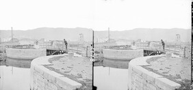 Navigation bridge (iron) on curved lock, dock on canal entrance, another bridge, towns old mill, hills, in background