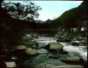Houses along bank of boulder-strewn  river; wooden bridge across river; stone embankment below houses; laundry hanging at edge of embankment.